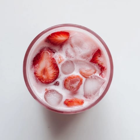 A tall glass of Refresher Copycat Pink Drink filled with ice, creamy coconut milk, and fresh sliced strawberries.  